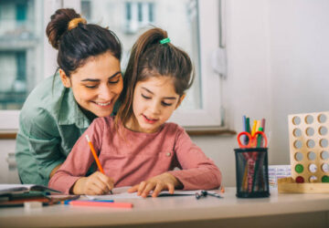 Une adulte souriante accompagne une fillette pendant qu'elle écrit à la table. Elles travaillent dans un environnement calme et chaleureux, avec des crayons colorés et du matériel éducatif. L’image évoque un moment d’apprentissage guidé et de complicité.