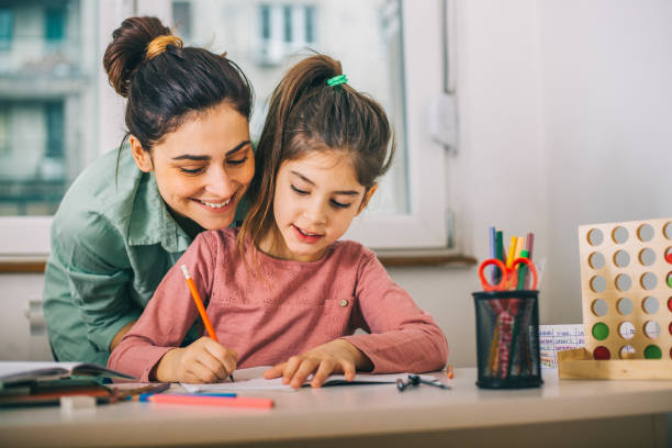 Une adulte souriante accompagne une fillette pendant qu'elle écrit à la table. Elles travaillent dans un environnement calme et chaleureux, avec des crayons colorés et du matériel éducatif. L’image évoque un moment d’apprentissage guidé et de complicité.