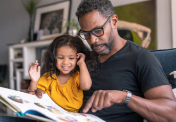 Un père lit un livre illustré avec sa jeune fille souriante assise sur ses genoux