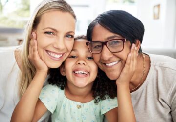 Enfant souriante entourée de deux adultes qui la tiennent par les joues, illustrant une famille chaleureuse et un environnement soutenant le développement du langage.